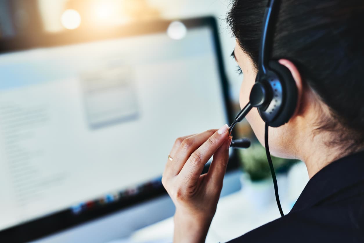 A woman wearing a phone headset sits at a computer looking at the screen while apparently talking to a customer. On the screen is a larger background window with information, and a small pop-out window.