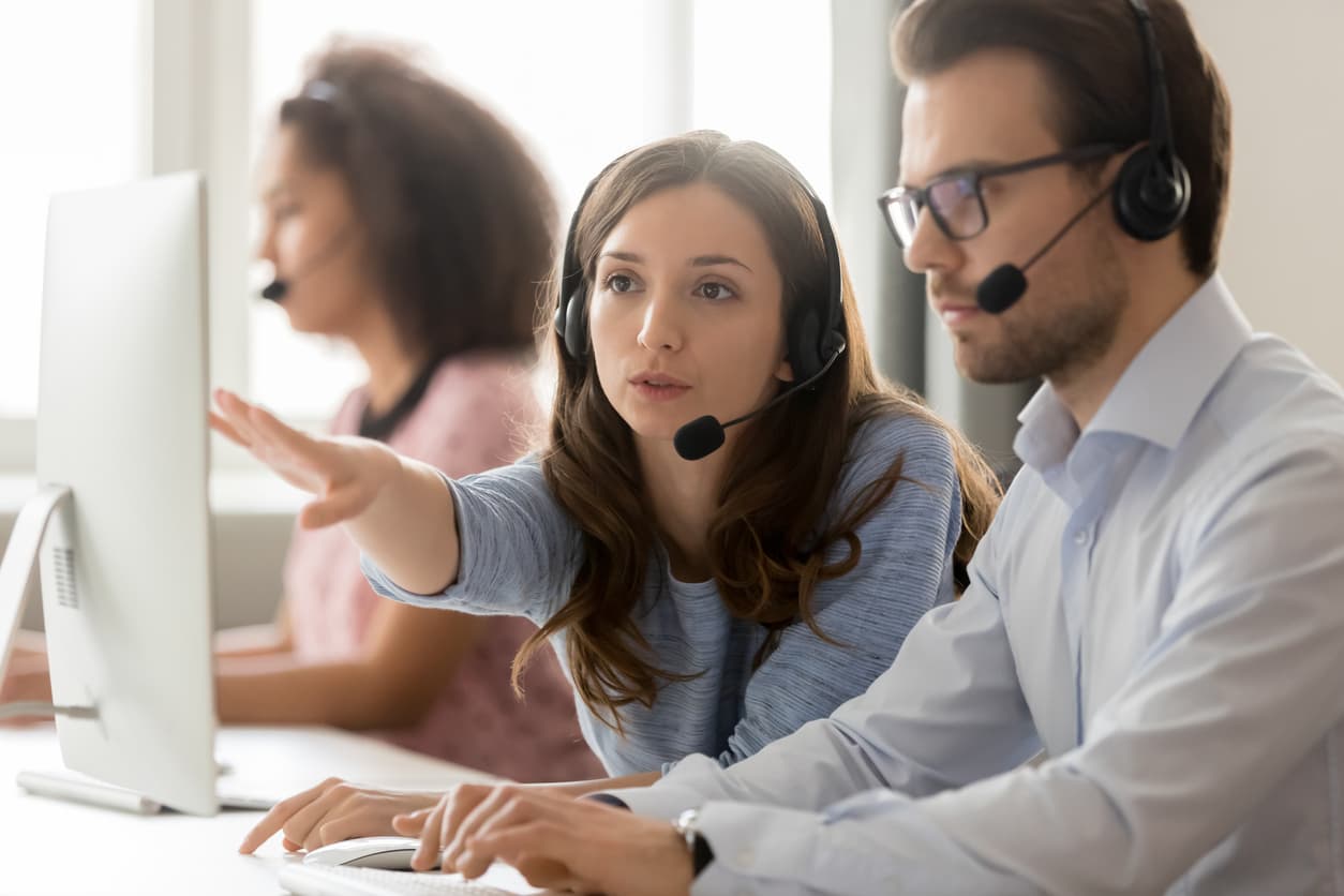 Set in a customer service call center, this image shows a female supervisor coaching a male customer service representative by talking while pointing at the computer screen. The customer service representative is typing as she speaks.