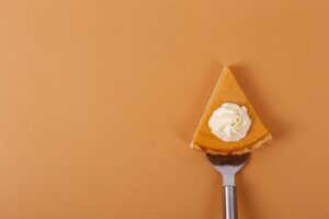 This image has an orange background. In the foreground is a slice of pie, also orange, with whipped cream on top sitting on a pie server. The hand holding the server is out of the frame. 