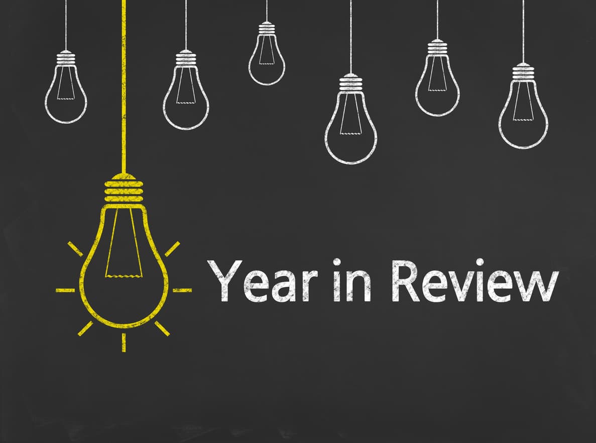 This image shows a blackboard with line drawings of lightbulbs in white, hanging at different levels from the top of the board. One lightbulb is in yellow, with rays coming off of it illustrating ideas, while letters say The Year in Review.