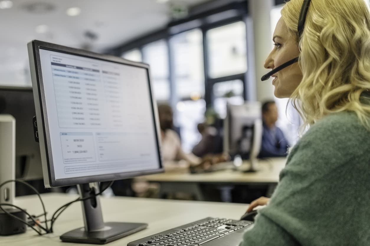 A female customer service agent wearing a headset and a green sweater sits at a desk looking at a computer screen.