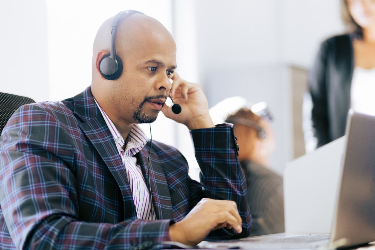 This image shows a man at a laptop computer sitting on a desk. He is looking at the screen while wearing an audio headset.