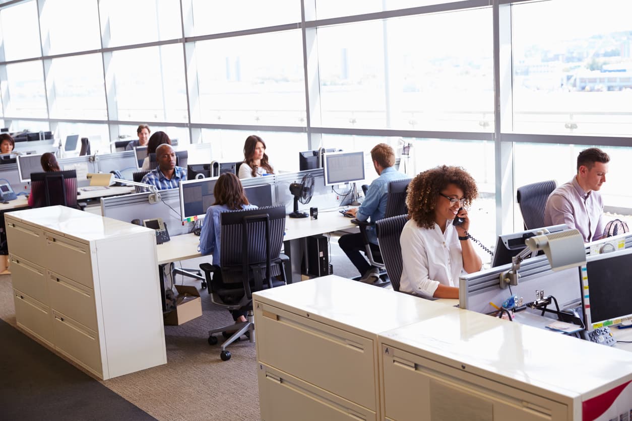 In a brightly-lit windowed room, workers sit at tables in front of computers.