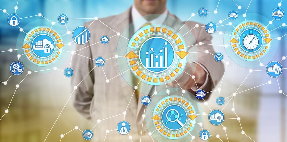 A man in a suit stands in front of a screen with outstretched arm. His finger is pointing at one of many descriptive circles on the screen such as search, profitability, security, connectivity, and others describing business technology.