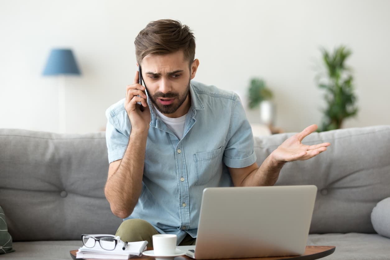 An agitated man sits on a couch in front of a laptop while talking to customer service over the phone.