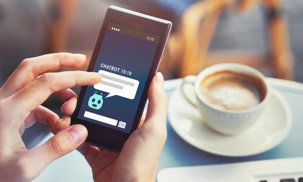 A person is using a chatbot on their mobile phone. They are sitting at a table that holds a laptop and a cappuccino.