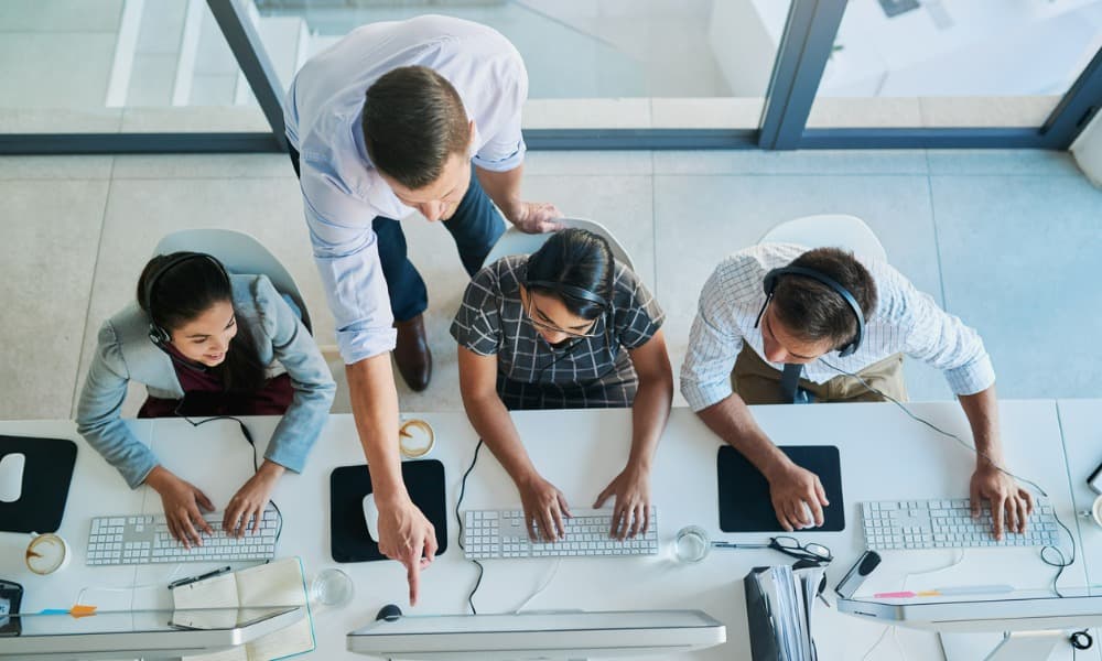 The image shows an overhead view of three customer service agents working at connected desks, with a supervisor providing tips or training in an office setting.