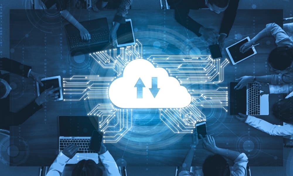 Against a turquoise background, workers sit around a table on their computers. In the center, a cloud has arrows that show data moving both ways, with information accessible by everyone present.